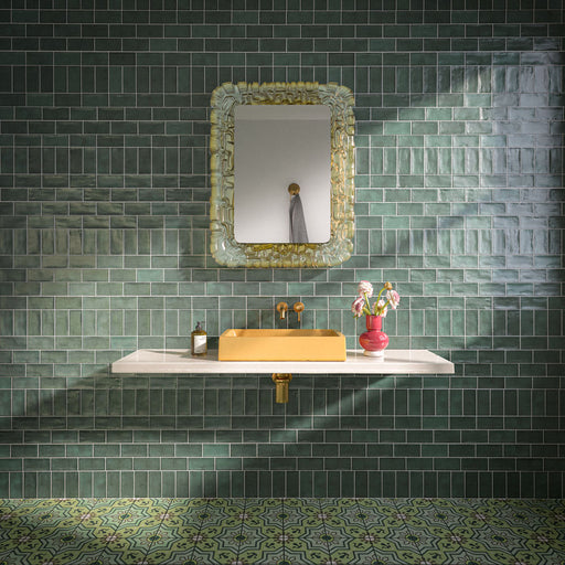 Bathroom featuring Dordogne Green Pines 7.5x15cm rustic gloss wall tiles, showcasing rich green tones with handmade-style shade variation, paired with a white floating vanity, mustard yellow basin, brass fittings, ornate mirror and patterned green floor tiles.