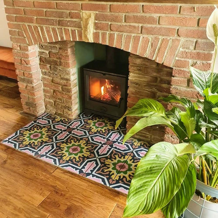 Fireplace hearth featuring Cuba Havana Moroccan-style porcelain tiles in a 20×20cm square format, showcasing a bold geometric pattern in navy, pink, green and mustard tones, framed by exposed brickwork and a wood-burning stove for a warm, character-filled finish.