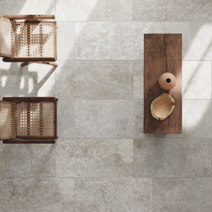 Flat lay of Flagstone Grey Tumbled Travertine 50 × 100cm porcelain floor tiles, styled with warm wood furniture and woven textures to highlight soft tonal variation, a gently aged finish, and a relaxed, natural stone effect.