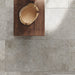 Flat lay of Flagstone Grey Tumbled Travertine 50 × 100cm porcelain floor tiles, styled with warm wood furniture and woven textures to highlight soft tonal variation, a gently aged finish, and a relaxed, natural stone effect.