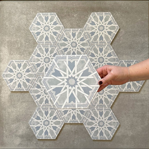 Hand holding Medina Hormuz Azure Blue Moroccan-style hexagon porcelain tile, 18.7 × 21.6 cm, shown against a grouped layout to highlight the bold star-inspired geometric pattern in soft blue and white tones.