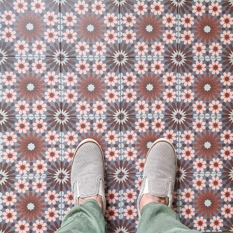 Fez Rosso Moroccan-style porcelain tiles laid in a neat grid, creating a rich, rhythmic pattern across the floor. Warm terracotta red starbursts sit against a soft grey-blue base, balanced by delicate blush pink and white geometric details inspired by traditional Moroccan tilework. Styled here with neutral footwear for scale, the design feels bold yet refined—perfect for adding warmth and character to kitchens, hallways or statement floor spaces while remaining practical and durable in porcelain.