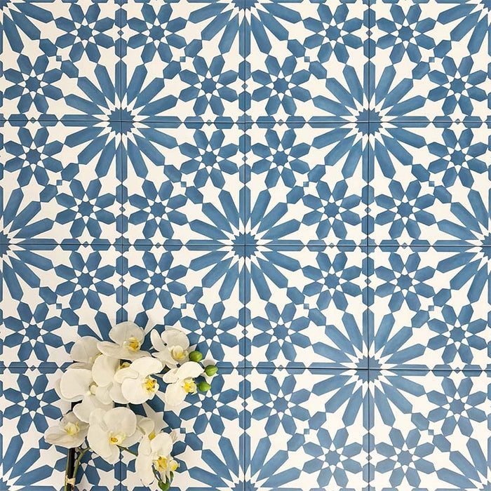 Fez Azure Blue 20x20cm Moroccan-style porcelain wall tiles arranged in a repeating geometric pattern, featuring crisp blue and white star motifs, styled with white orchid flowers for a bright, decorative finish.