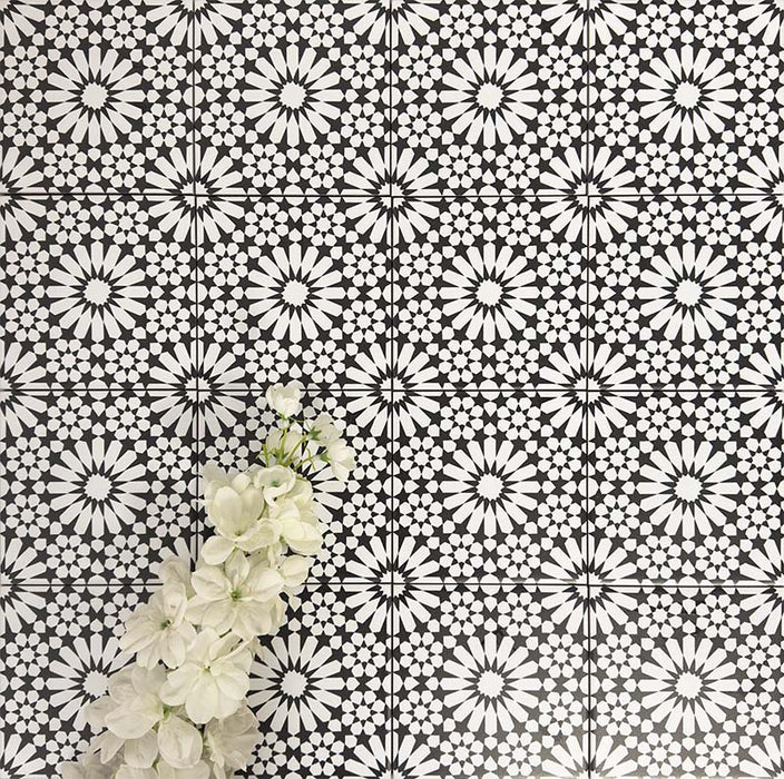 Fez Mini Winter Moroccan-style porcelain tiles shown laid in a repeating pattern, featuring a striking black and white geometric star design inspired by traditional Moroccan motifs. The 15 × 15cm tiles create a bold, high-contrast surface with intricate detailing, styled here with white flowers for a clean, elegant look—ideal for statement floors, splashbacks, or feature walls in both modern and classic interiors.