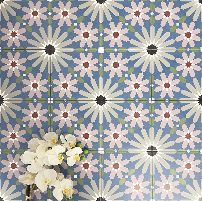 Fez Artisan 20x20cm Moroccan-style porcelain wall tiles arranged in a repeating geometric pattern, featuring soft blue, blush pink, green and terracotta star motifs, styled with white orchid flowers for a warm, decorative finish.