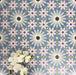 Fez Artisan 20x20cm Moroccan-style porcelain wall tiles arranged in a repeating geometric pattern, featuring soft blue, blush pink, green and terracotta star motifs, styled with white orchid flowers for a warm, decorative finish.