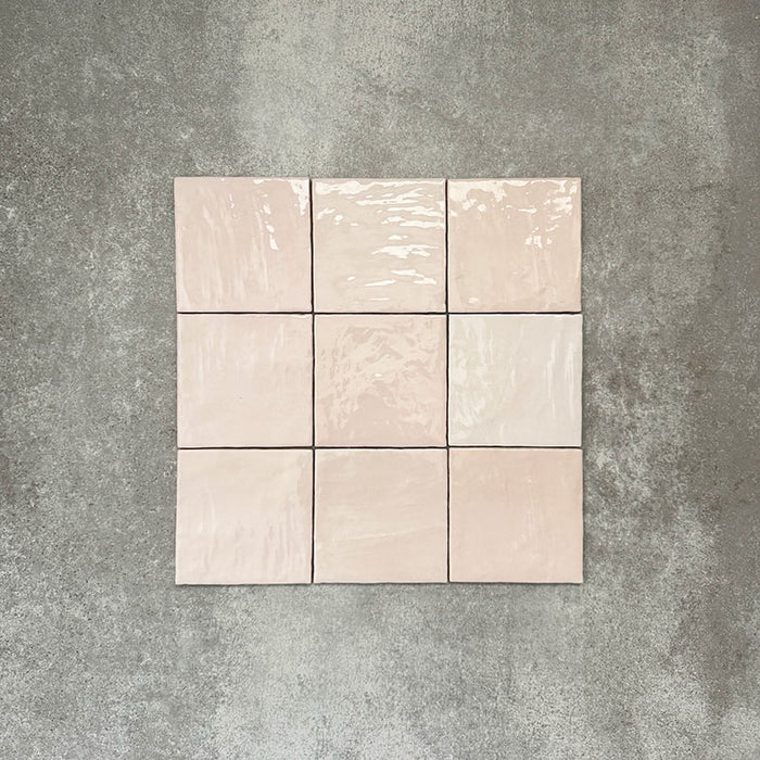 Flat lay of Zellige Pink Mist Square 13.2 × 13.2cm Moroccan-style ceramic tiles in soft pale pink and blush tones, arranged in a simple grid on a textured grey background, showcasing the glossy glaze, subtle tonal variation and handcrafted zellige-effect finish.