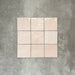 Flat lay of Zellige Pink Mist Square 13.2 × 13.2cm Moroccan-style ceramic tiles in soft pale pink and blush tones, arranged in a simple grid on a textured grey background, showcasing the glossy glaze, subtle tonal variation and handcrafted zellige-effect finish.