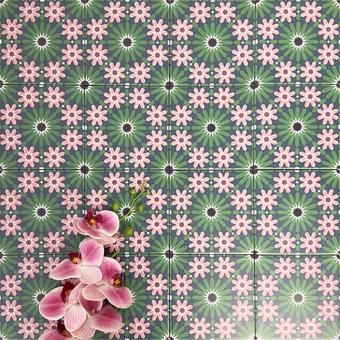 Fez Mini Autumn Moroccan-style porcelain tiles shown laid in a repeating pattern, creating a richly detailed geometric design. The 15 × 15cm tiles feature soft blush pink floral motifs, deep green starbursts and charcoal accents on a muted blue-grey background. Styled with pink orchids, the image highlights the encaustic-effect finish and the decorative impact of this pattern when used across a larger surface.