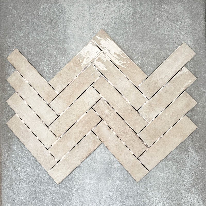 Nomad Oatmeal brick wall tiles in a 6 × 24.6cm format, shown laid together in a herringbone layout to highlight the warm neutral tone, gloss finish and subtle surface variation.