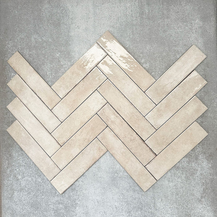 Nomad Oatmeal brick wall tiles in a 6 × 24.6cm format, shown laid together in a herringbone layout to highlight the warm neutral tone, gloss finish and subtle surface variation.