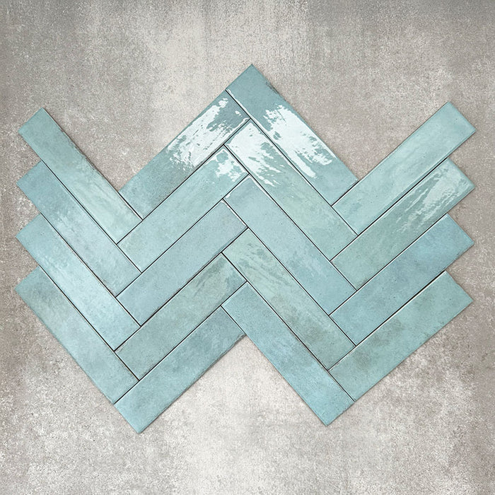 Nomad Aquamarine brick wall tiles in a 6 × 24.6cm format, shown laid in a herringbone layout with a glossy, lightly textured surface and soft blue-green tones, creating an industrial-style look.