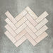Flat lay of Zellige Pink Mist Brick 6.5 × 20cm Moroccan-style ceramic tiles in soft pale pink and blush tones, arranged in a herringbone layout on a textured grey background, showcasing the glossy glaze, gentle tonal variation and handcrafted zellige-effect finish.