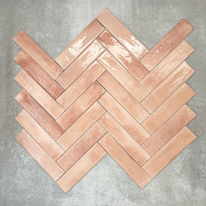Allegra Tusk Pink Gloss Brick ceramic tiles in a 6 × 25cm format, arranged in a herringbone pattern to showcase the soft pink tones, glossy finish and subtle handmade-style variation.