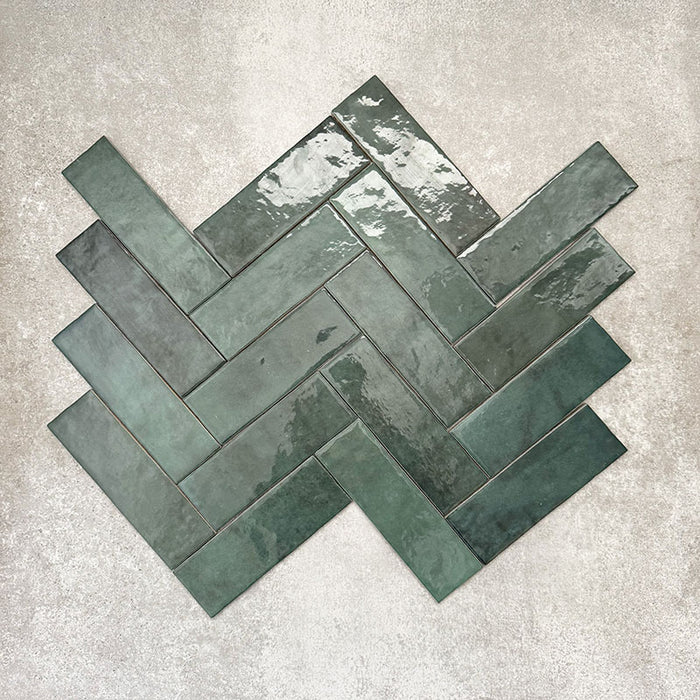 Flat lay of Zellige Royal Green Brick 6.5 × 20cm Moroccan-style ceramic tiles in rich deep green tones, arranged in a herringbone layout on a textured grey background, showcasing the glossy glaze, natural tonal variation and handcrafted zellige-effect finish.