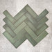 Flat lay of Zellige Malachite Green 6.5×20cm Moroccan-style ceramic brick tiles, arranged in a herringbone layout, showcasing subtle tonal variation, handmade-look texture and rich green glaze on a neutral background.
