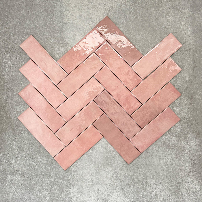 Flat lay of Zellige Rose Pink Brick 6.5 × 20cm Moroccan-style ceramic tiles, shown in soft blush, dusty rose and terracotta pink tones with gentle tonal variation and lightly uneven edges, arranged in a herringbone brick layout on a textured grey background to showcase the handcrafted zellige-effect glaze.