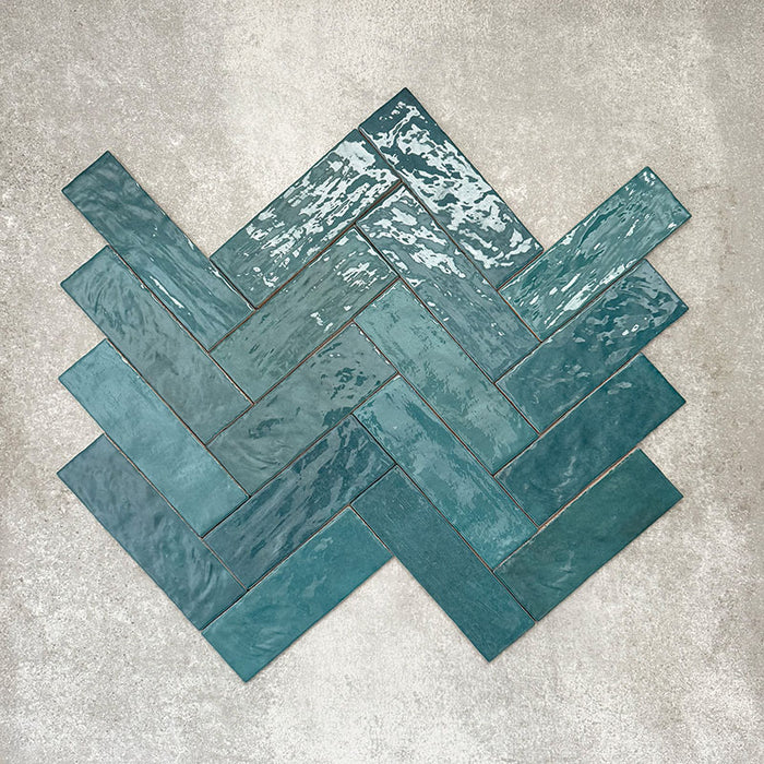 Flat lay of Zellige Peacock Green 6.5×20cm Moroccan-style ceramic brick tiles, shown in a herringbone layout, showcasing deep blue-green tones, a glossy handmade-look glaze and natural tonal variation on a neutral background.