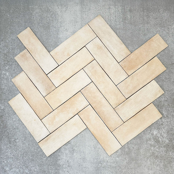 Flat lay of Zellige Arena Marfil Brick 6.5 × 20cm Moroccan-style ceramic tiles, shown in a warm beige and cream colour palette with subtle tonal variation and gently uneven edges, arranged in a herringbone layout on a textured grey background to highlight the handcrafted, zellige-effect finish.