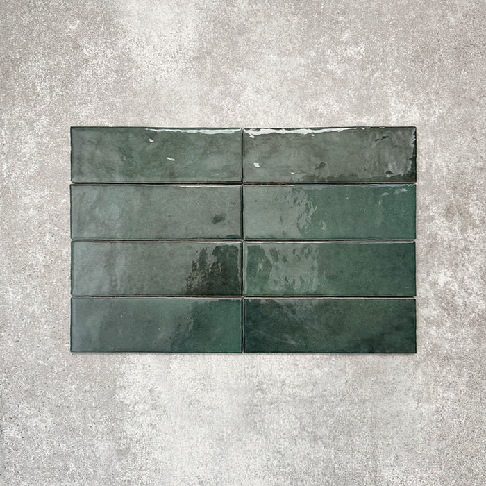 Flat lay of Zellige Royal Green Brick 6.5 × 20cm Moroccan-style ceramic tiles in rich deep green tones, arranged in a horizontal brick layout on a textured grey background, showcasing the glossy glaze, natural tonal variation and handcrafted zellige-effect finish.