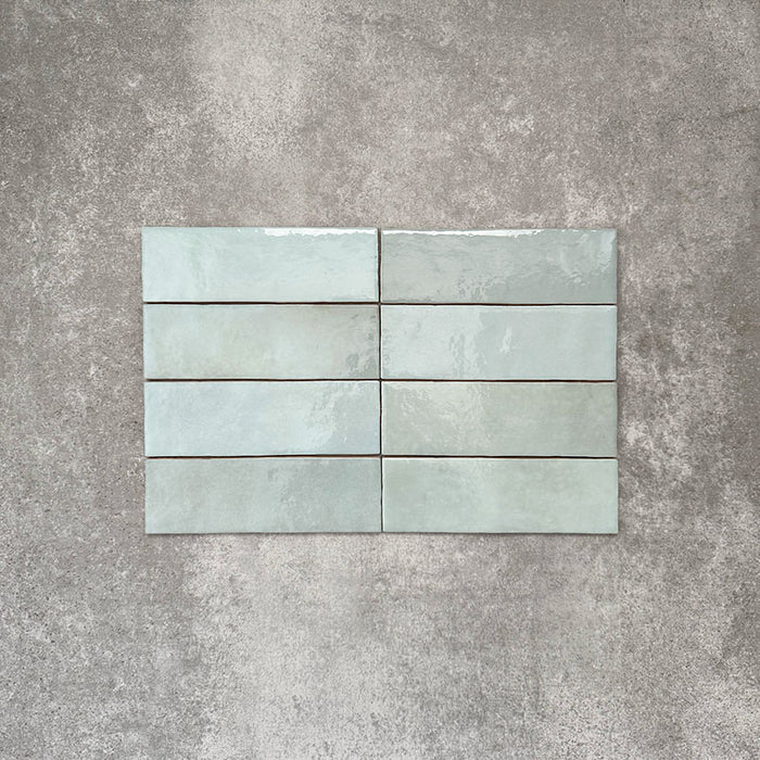Flat lay of Zellige Aqua 6.5×20cm Moroccan-style ceramic brick tiles, showcasing soft blue-green tones, a glossy handmade-look glaze and subtle tonal variation on a neutral background.