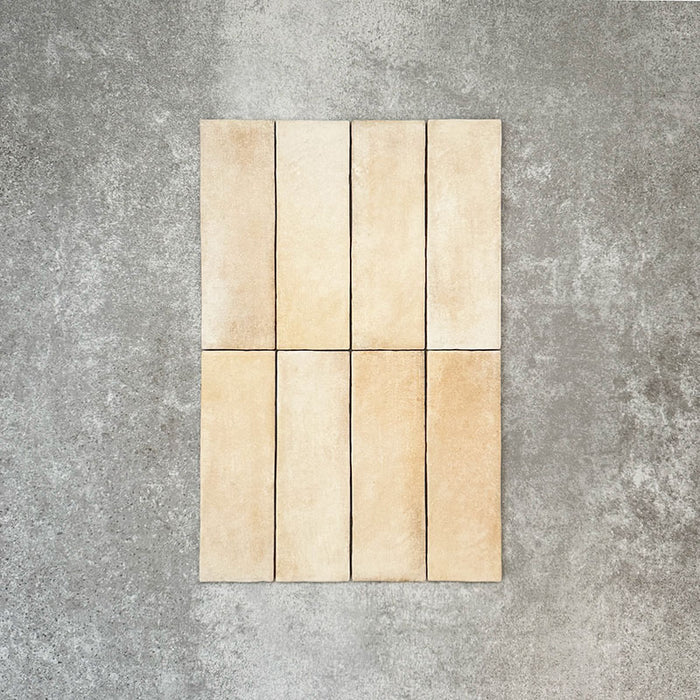 Flat lay of Zellige Arena Marfil Brick 6.5 × 20cm Moroccan-style ceramic tiles, shown in a warm beige and cream colour palette with subtle tonal variation and gently uneven edges, arranged in a vertical brick layout on a textured grey background to highlight the handcrafted, zellige-effect finish.