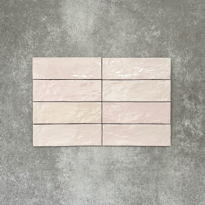 Flat lay of Zellige Pink Mist Brick 6.5 × 20cm Moroccan-style ceramic tiles in soft pale pink and blush tones, arranged in a horizontal brick layout on a textured grey background, showcasing the glossy glaze, gentle tonal variation and handcrafted zellige-effect finish.