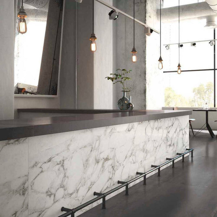 Bar and dining setting featuring Hudson Calacatta Matt Marble 30 × 60cm porcelain wall tiles, styled across the counter front to showcase soft grey veining on a clean white base, paired with dark worktops, industrial lighting, and polished concrete for a refined, contemporary contrast.