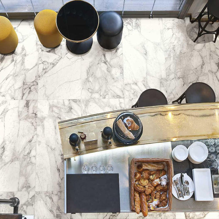 Top-down view of Hudson Calacatta Matt Marble 60 × 60cm porcelain floor tiles, showcasing a soft white base with elegant grey veining, styled within a café setting with warm metallic accents and contemporary furnishings for a refined, modern look.