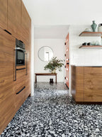 Kitchen setting featuring Shards Terrazzo Nero 59x118cm matt porcelain wall and floor tiles, showcasing a bold black base with contrasting white aggregate, paired with warm wood cabinetry, open shelving and a clean, contemporary finish.