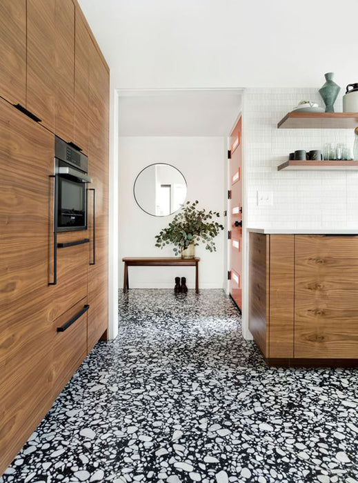 Kitchen setting featuring Shards Terrazzo Nero 59x118cm matt porcelain wall and floor tiles, showcasing a bold black base with contrasting white aggregate, paired with warm wood cabinetry, open shelving and a clean, contemporary finish.
