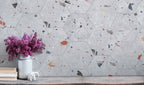Styled interior wall featuring Jericho Silver Terrazzo Hexagon 25 × 22cm porcelain tiles, with a soft grey base and scattered multicoloured aggregate chips, laid in a honeycomb pattern and paired with natural wood, books, and floral accents for a relaxed, contemporary look.