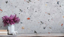 Styled interior wall featuring Jericho Silver Terrazzo Hexagon 25 × 22cm porcelain tiles, with a soft grey base and scattered multicoloured aggregate chips, laid in a honeycomb pattern and paired with natural wood, books, and floral accents for a relaxed, contemporary look.