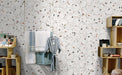 Retail-style interior wall featuring Jericho White Terrazzo Hexagon 25 × 22cm porcelain tiles, with a crisp white base and scattered multicoloured aggregate chips, laid in a honeycomb pattern and paired with light wood shelving and soft textiles for a fresh, contemporary display.