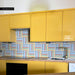 Kitchen interior featuring Pinstripes Rainbow 20x20cm patterned tiles installed as a colourful splashback, styled with bold yellow cabinetry, wood worktops, and brass tap for a playful, contemporary design.
