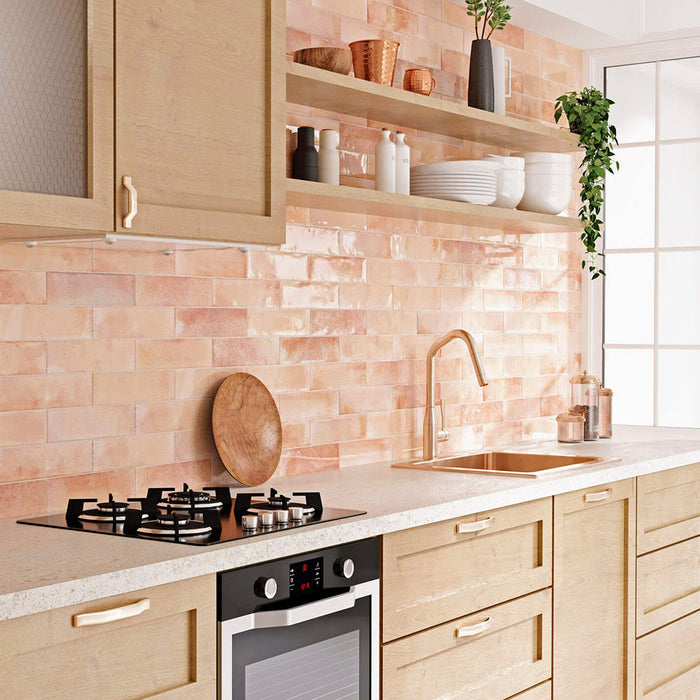 Kitchen wall splashback featuring Allegra Tusk Pink Gloss Brick ceramic tiles in a 6 × 25cm format, styled with warm wood cabinetry and open shelving to highlight the soft pink tones, glossy finish and handmade-style character.