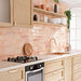 Kitchen wall splashback featuring Allegra Tusk Pink Gloss Brick ceramic tiles in a 6 × 25cm format, styled with warm wood cabinetry and open shelving to highlight the soft pink tones, glossy finish and handmade-style character.