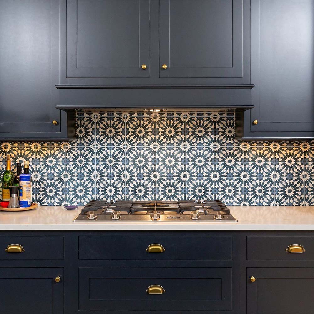 Kitchen splashback featuring Fez Federal Blue 20x20cm Moroccan-style porcelain tiles, showcasing a repeating blue and white geometric star pattern, paired with dark navy cabinetry, brass handles and a modern stainless steel hob for a bold, elegant finish.