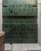 Walk-in shower featuring Levent Green 6 × 24cm porcelain wall tiles, laid vertically with rich green tonal variation and a glossy, handcrafted look, paired with dark shower fittings, a recessed shelf, and light stone-effect flooring for contrast.