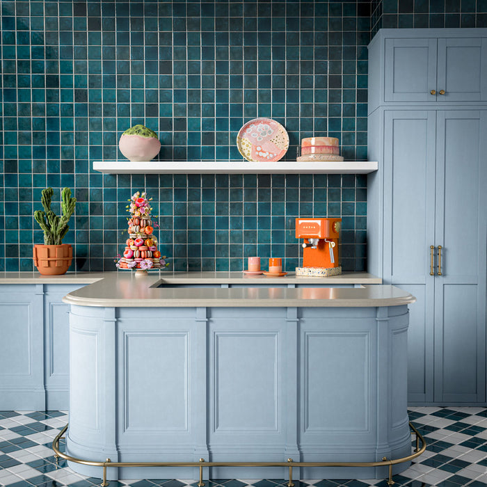 Kitchen splashback featuring Levent Blue Square 10cm x 10cm gloss porcelain wall tiles, showcasing rich blue tones with subtle handmade-effect shade variation, paired with pale blue cabinetry, light stone worktops, open shelving and vibrant contemporary accessories.