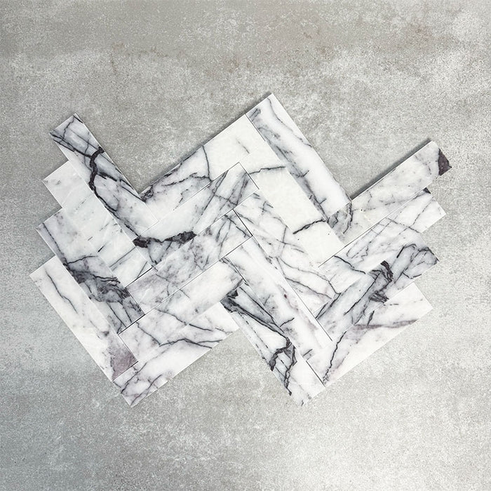 Arrangement of Fayrouz Lilac 5x20cm natural marble tiles with a honed finish, displayed in a herringbone layout, showcasing a soft white base with flowing lilac and grey veining, highlighting natural variation and adding movement and elegance to the design.