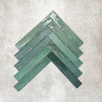 Close-up of Lisbon Verde Green 5cm x 30cm artisanal porcelain wall tiles arranged in a herringbone pattern, showcasing rich green tones, subtle tonal variation and a handcrafted-effect surface finish.