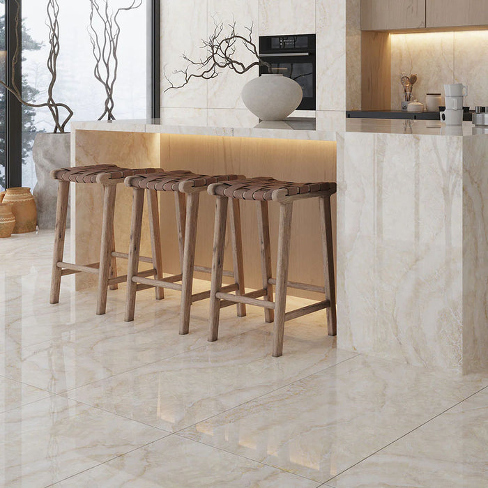 Open-plan kitchen interior featuring Lumen Precious Stone Ivory 60 × 120cm polished porcelain wall and floor tiles, showcasing warm ivory and cream gemstone-inspired tones with crystalline layering, delicate natural veining, and a reflective high-gloss finish, paired with light wood cabinetry, a statement island, woven leather bar stools, and soft ambient lighting.