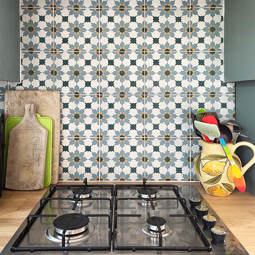 Madeira Anza 20cm x 20cm Moroccan style wall and floor tiles, featuring an intricate geometric pattern in blue, yellow and white, creating a colourful kitchen splashback paired with muted blue cabinetry and warm wooden worktops.