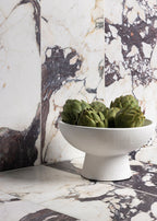 Interior setting featuring Fayrouz Viola 30.5x61cm natural marble tiles with a honed finish, installed across walls and surfaces, showcasing a white base with bold burgundy and warm-toned veining, styled with a decorative bowl and accessories.
