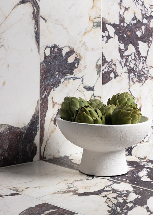 Interior setting featuring Fayrouz Viola 30.5x61cm natural marble tiles with a honed finish, installed across walls and surfaces, showcasing a white base with bold burgundy and warm-toned veining, styled with a decorative bowl and accessories.