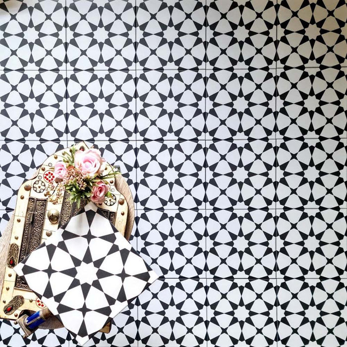 Marketing image featuring Agadir Black & White Moroccan-style 20x20cm porcelain floor tiles laid in a repeating geometric star pattern, styled with decorative accessories to highlight the bold black and white design.