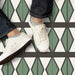 Abstract Hourglass Patterned Tiles in a 20x20cm format, laid as a geometric floor design featuring an hourglass motif in muted green and soft cream tones with charcoal detailing. Styled with casual footwear to highlight durability and pattern repetition, making the tiles ideal for kitchens, hallways or high-traffic interior spaces.
