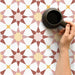 Marketing image featuring Agadir Springs Moroccan-style 20x20cm porcelain floor tiles laid in a repeating geometric star pattern, showcasing soft terracotta, blush pink, white and warm yellow tones, styled with a coffee cup to highlight everyday use.