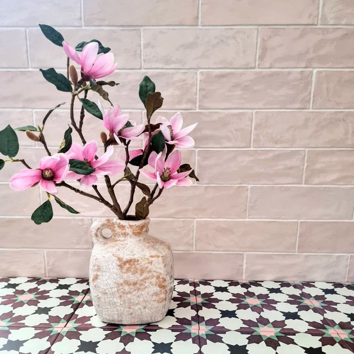 Interior detail featuring Zellige Pink Mist Brick 6.5 × 20cm Moroccan-style ceramic wall tiles in soft pale pink tones, laid in a horizontal brick layout behind a decorative vase and patterned tiles, highlighting the glossy surface, gentle colour variation and handcrafted zellige-effect finish.